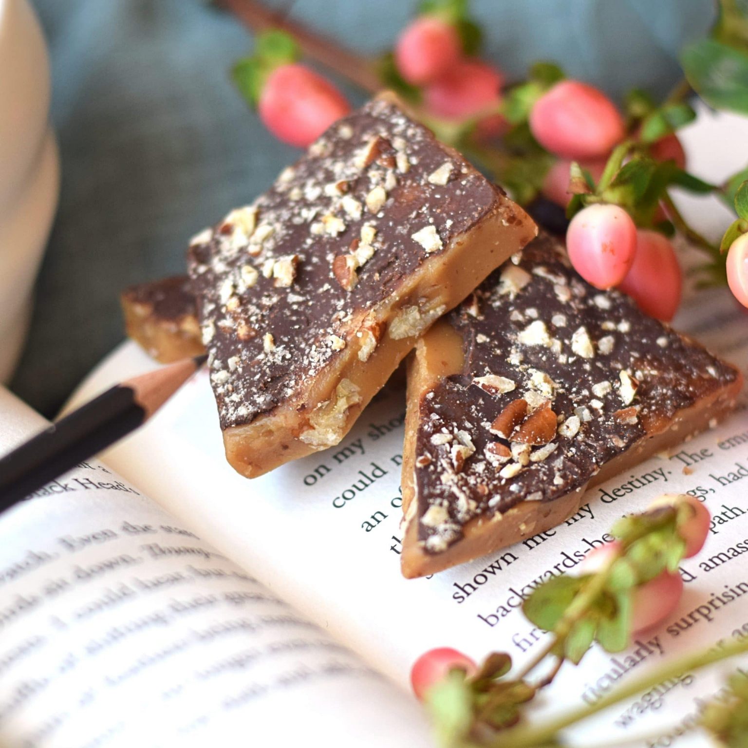 Stack of pieces of English toffee with gourmet dark chocolate spread on top and then sprinkled with chopped pecans; the pieces are sitting on an open book with flower buds in the back and foregrounds