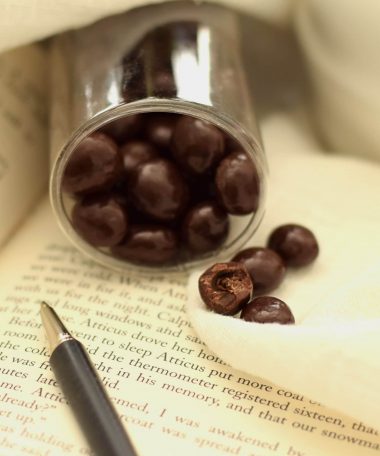 4 espresso beans covered in gourmet dark chocolate spilling out of an open package. Items are sitting on an open book; one piece has a bite taken out of it