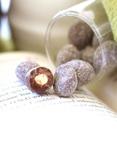 4 almonds covered in toffee and gourmet milk chocolate and sprinkled with powdered sugar spilling out of an open package. Items are sitting on an open book; one piece has a bite taken out of it