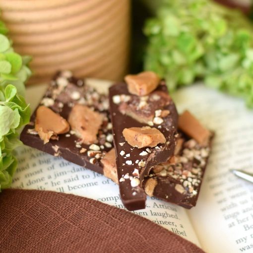 Stack of 2 pieces of an artisan chocolate bar sitting on an open book; the bar has toffee pieces on top and inside of a gourmet dark chocolate artisan bar