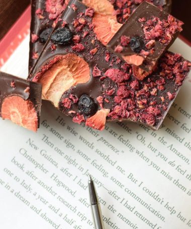 Pieces of a gourmet dark chocolate artisan bar embedded with dried raspberries, blueberries, and strawberries sitting on a pink book