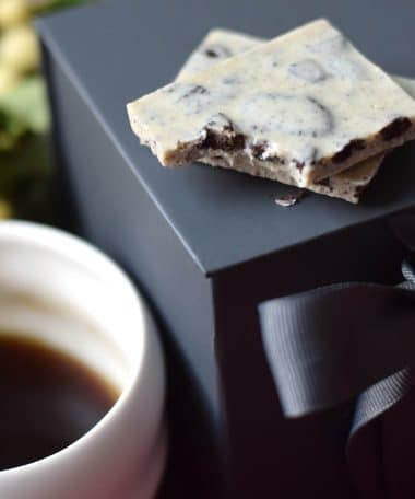 Two pieces of a gourmet white chocolate bar embedded with crushed Oreos on a box next to a cup of coffee
