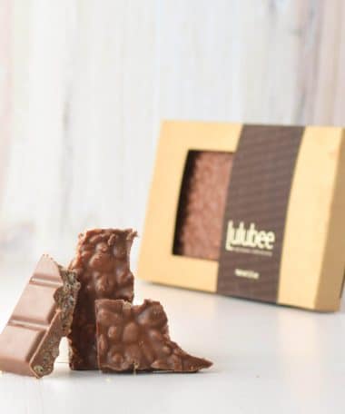 Three pieces of a gourmet milk chocolate bar embedded with puffed rice cereal in front of packaging
