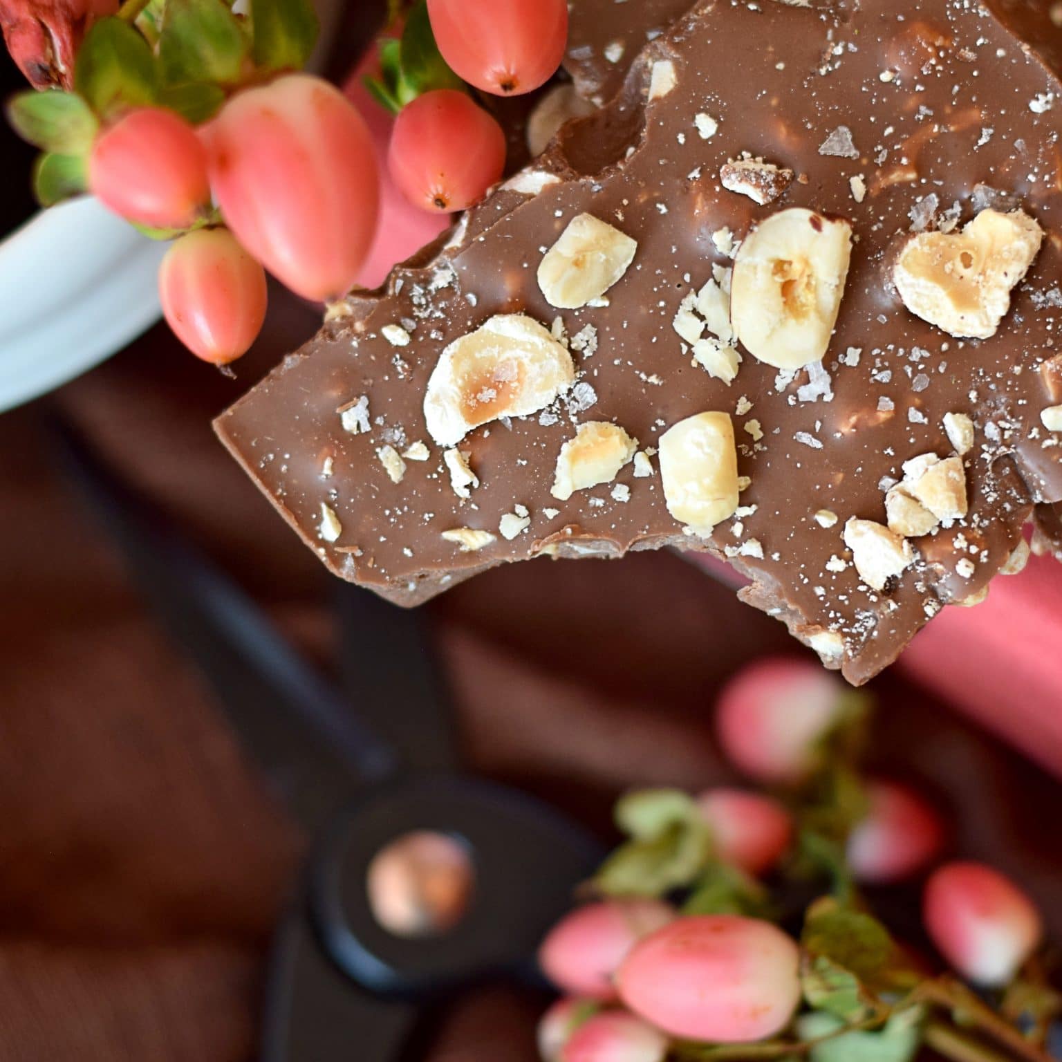 Piece of an artisan chocolate bar sitting on an pink book with pink flowers surrounding the bar. The milk chocolate bar is topped with pieces of salted hazelnuts