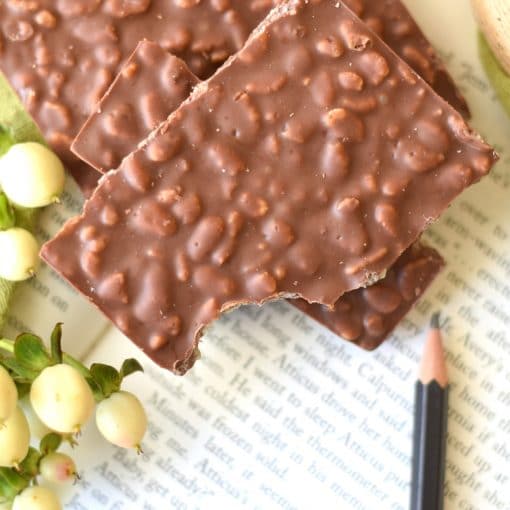 Stack of 2 pieces of an artisan chocolate bar sitting on an open book; the bar is embedded with crispy rice cereal and is made using gourmet milk chocolate
