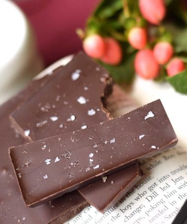 Stack of 3 pieces of an artisan chocolate bar sitting on an open book; the bar is sprinkled with sea salt and is made using gourmet dark chocolate