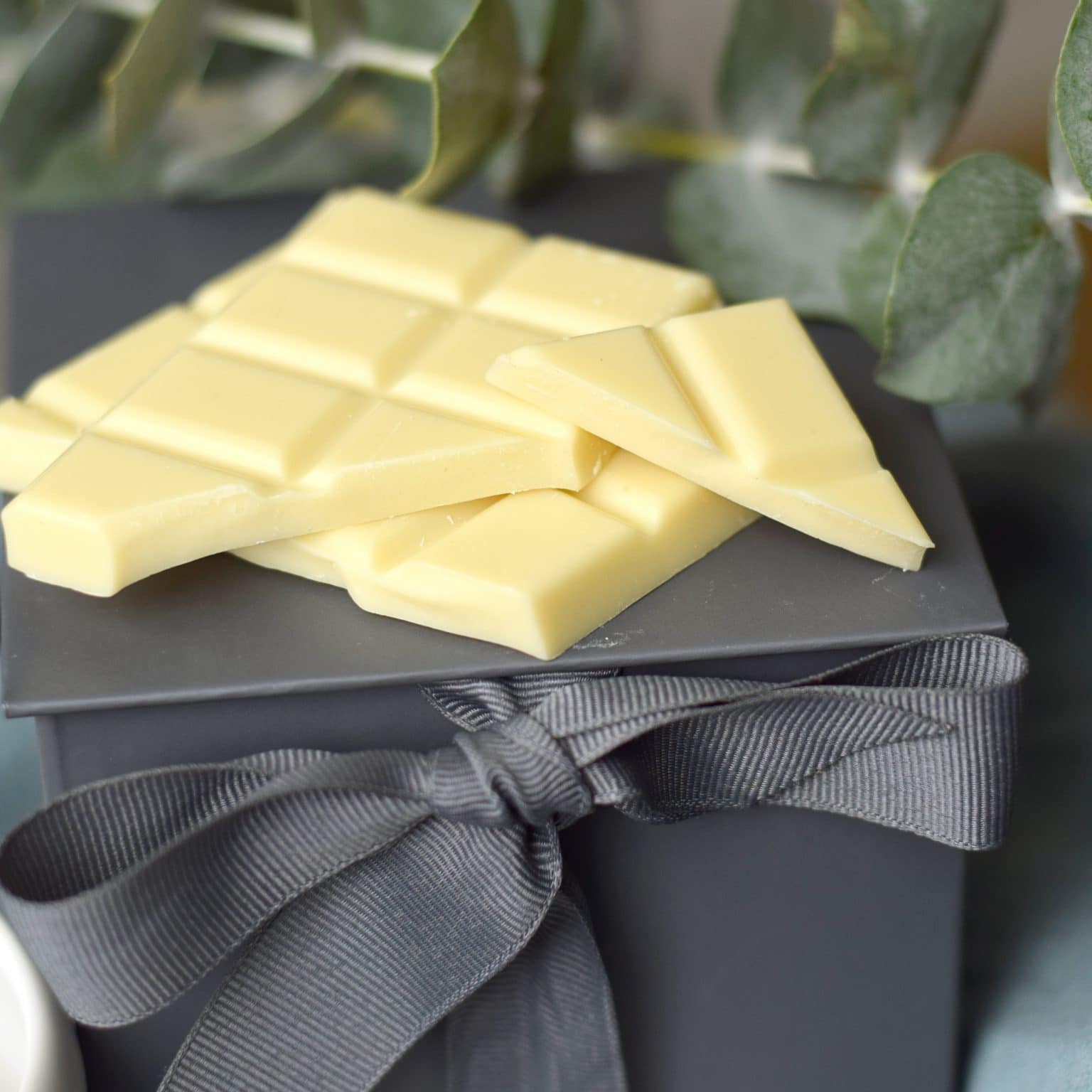 Stack of 3 pieces of an artisan chocolate bar sitting on gray gift box with a bow; bar is made with gourmet white chocolate