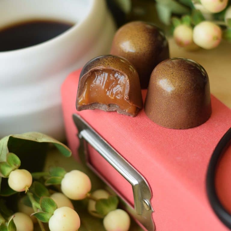 3 gourmet chocolate truffles sitting on a pink book with a metal label holder on the spine. The 3 truffles are light brown. 1 truffle has a bite out of it and browned butter caramel is dripping out