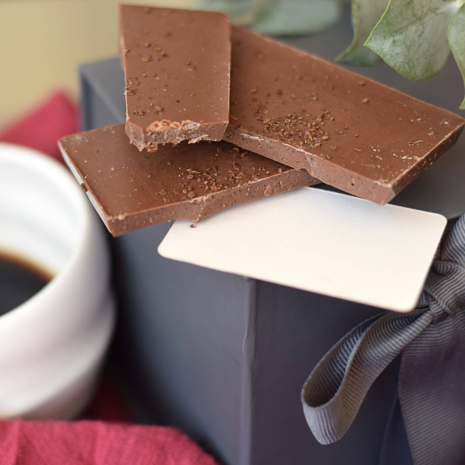 Stack of 3 pieces of a brown artisan chocolate bar sitting on a gray gift box and a white tag; bar has bits of espresso on top; made with gourmet milk chocolate