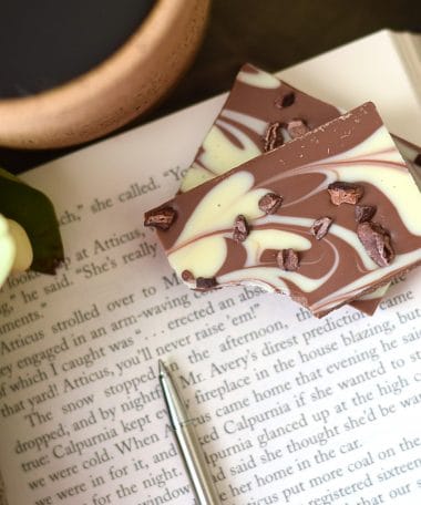 Stack of 2 pieces of an artisan chocolate bar sitting on an open book; the bar has swirls of milk and white gourmet chocolate and has a sprinkle of cacao nibs