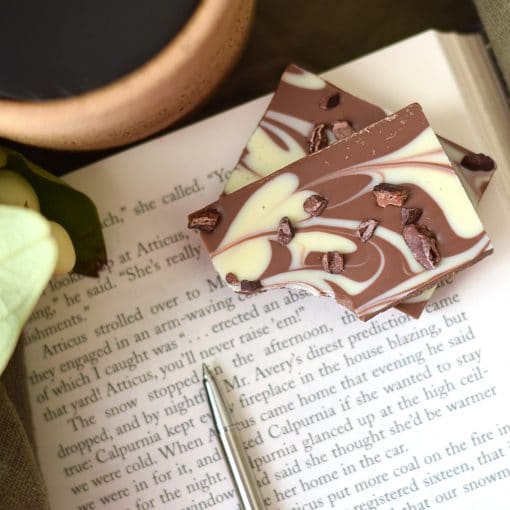 Stack of 2 pieces of an artisan chocolate bar sitting on an open book; the bar has swirls of milk and white gourmet chocolate and has a sprinkle of cacao nibs