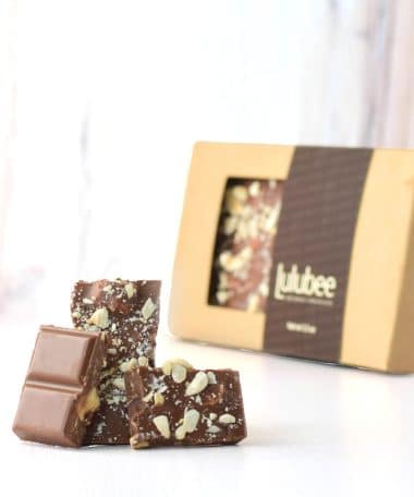 Three pieces of a gourmet milk chocolate bar embedded with salted hazelnuts in front of packaging