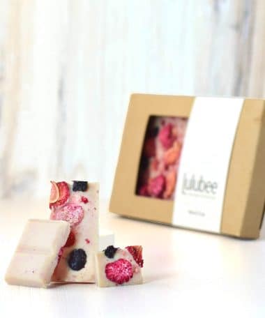 Three pieces of a gourmet white chocolate bar embedded with dried raspberries, blueberries, and strawberries in front of packaging