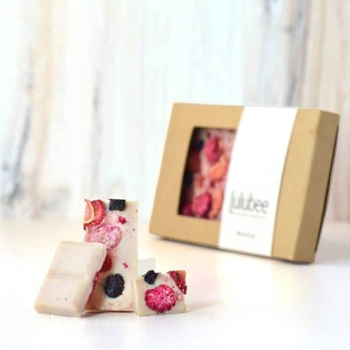 Three pieces of a gourmet white chocolate bar embedded with dried raspberries, blueberries, and strawberries in front of packaging