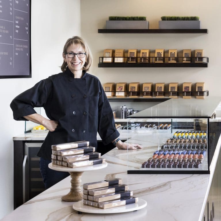 Smiling person in a store with gourmet chocolate truffles on the counter and in a glass-enclosed display. 3 shelves with brown package are in the background