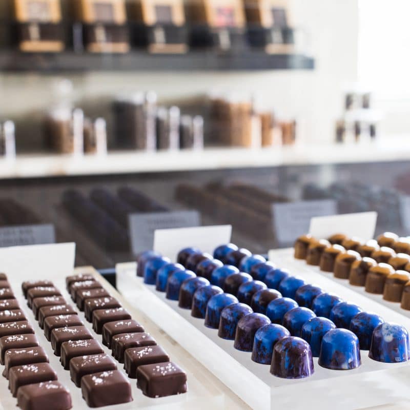 3 trays with 3 rows of gourmet chocolate truffles on each one. The truffles on the left are dark brown with sea salt sprinkled on them, dark blue in the center, and tan the right. Shelving with product packaging is in the background