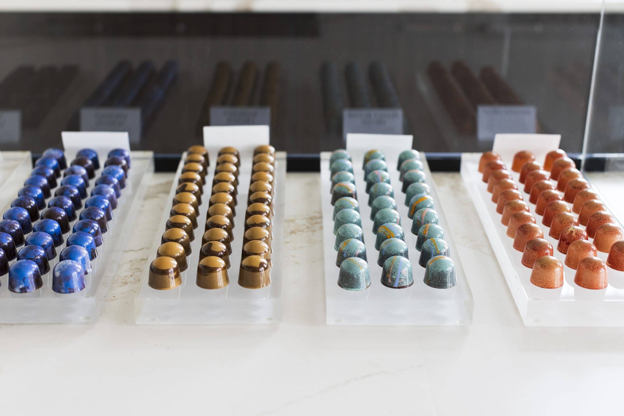 4 trays with 3 rows of gourmet chocolate truffles on each one. The truffles on the left are dark blue, then tan, light blue, and orange on the on the right