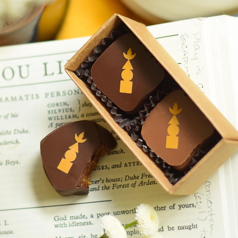 3 gourmet chocolate truffles, 2 in a box, sitting on an open book that contains a vertical, geometric-shaped logo on each one