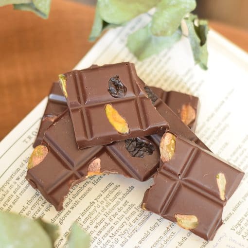 Pieces of a gourmet dark chocolate bar with pistachios and dried cherries embedded in the chocolate. Pieces are sitting on an open book