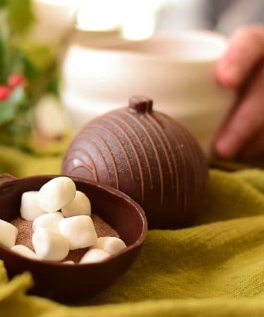 2 gourmet dark chocolate Christmas ornament-shaped balls. One is in half and filled with cocoa mix and marshmallows. A person taking a mug is in the background.