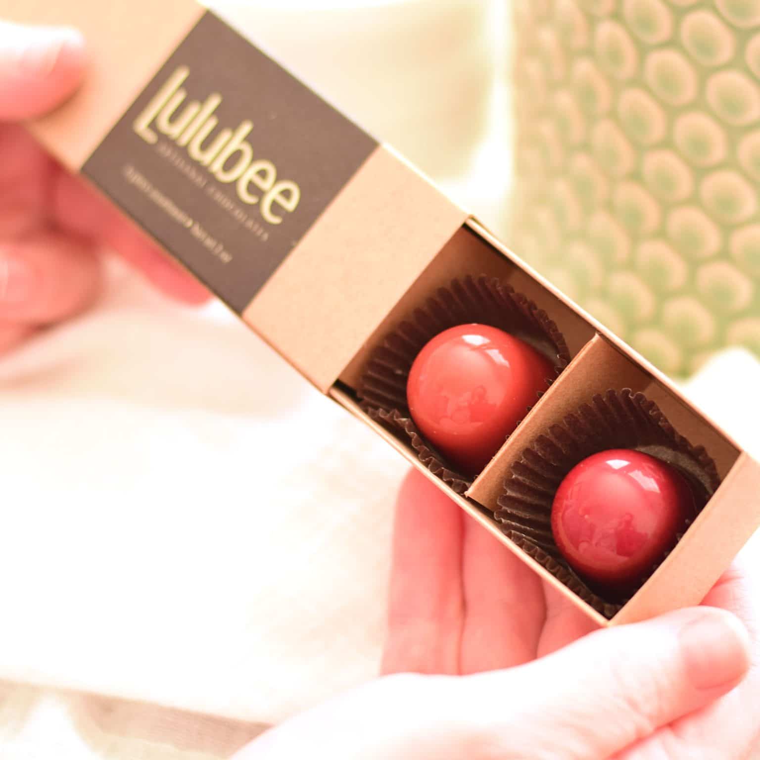 Person holding a 6-piece box of gourmet chocolate truffles in shades of red. Box contains a printed label that reads Lulubee Artisanal Chocolates