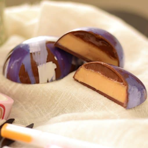 2 hand-painted large gourmet milk chocolate Easter eggs. One egg is cut and is filled with a thick layer of handmade peanut butter and a thin layer of caramel