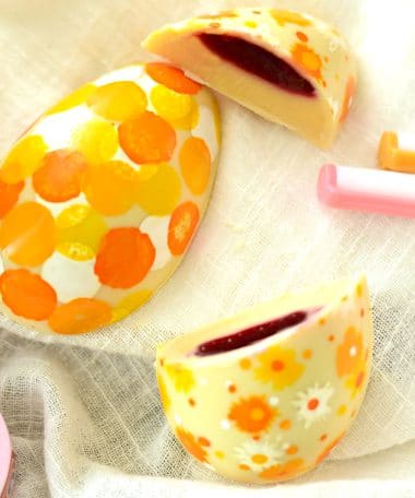 2 hand-painted large gourmet white chocolate Easter eggs. One egg is cut and is filled with a thick layer of lemon ganache and a thin layer of raspberry jelly