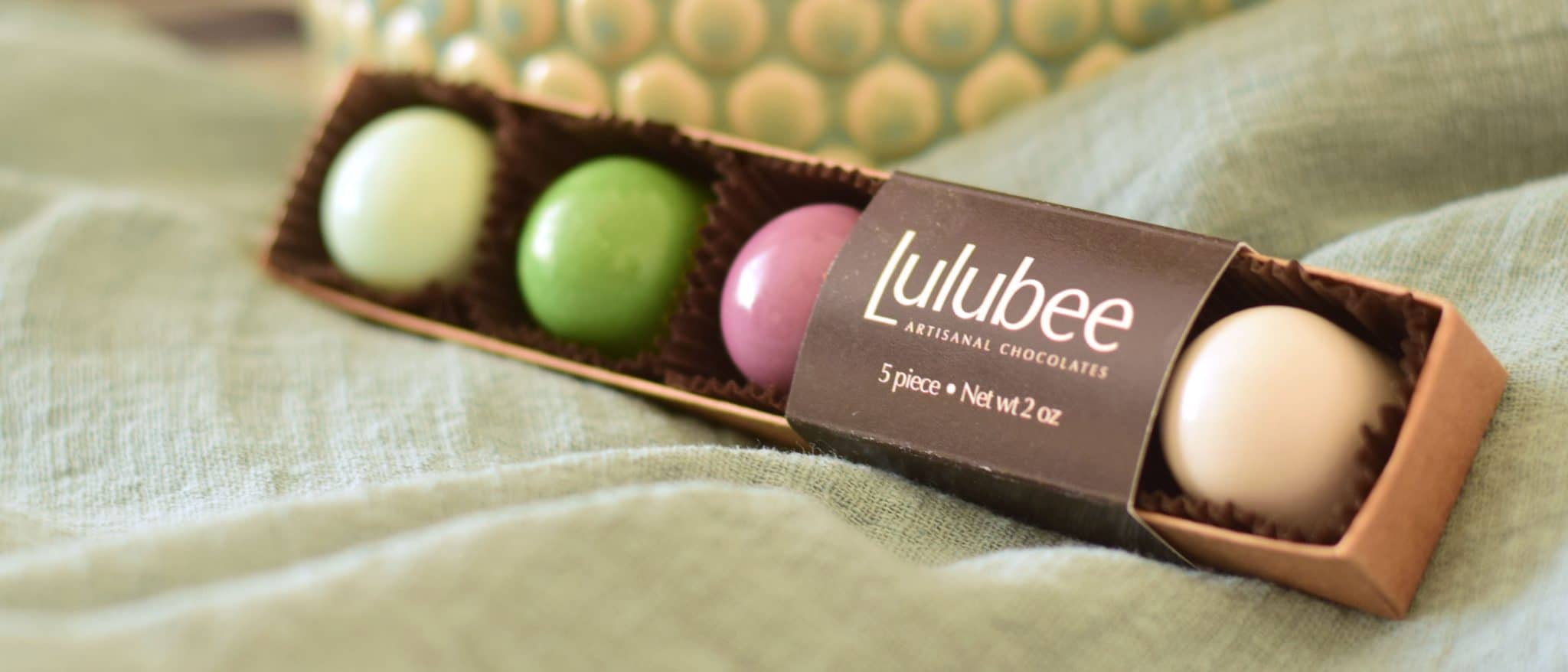 5 gourmet chocolate, pastel-colored truffles in a box that has a cigar band that reads Lulubee Artisanal Chocolates