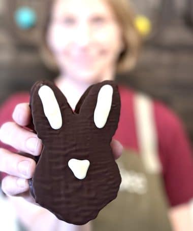 Person, who is out of focus, holding an Easter bunny-shaped head that is covered in gourmet dark chocolate and filled with vanilla bean marshmallow