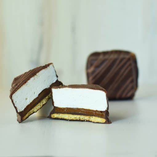 A gourmet s’more cut in half with a whole s’more in the background; s’more has a layer of graham cracker, caramel, and vanilla bean marshmallow covered in dark chocolate