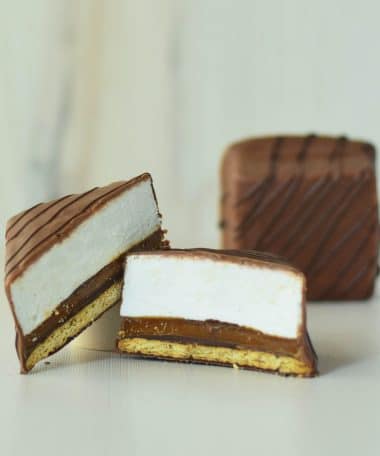 A gourmet s’more cut in half with a whole s’more in the background; s’more has a layer of graham cracker, caramel, and vanilla bean marshmallow covered in milk chocolate