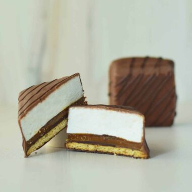 A gourmet s’more cut in half with a whole s’more in the background; s’more has a layer of graham cracker, caramel, and vanilla bean marshmallow covered in milk chocolate