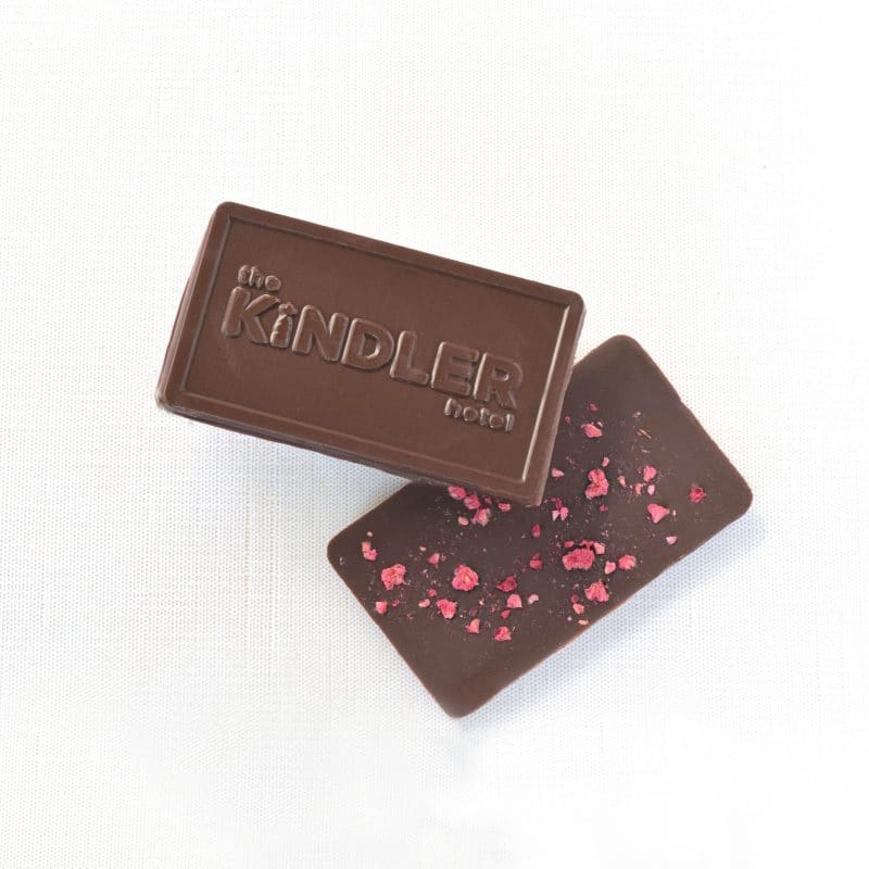 Front and back of a dark chocolate mini bar; one side has a company's logo embossed on it and the other is sprinkled with bits of dried raspberry