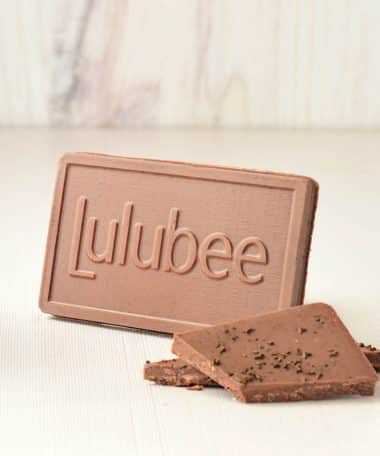 Small milk chocolate bar with the Lulubee logo on it; small bits of the chocolate bar are in front of the full bar. The bar is sprinkled with espresso.